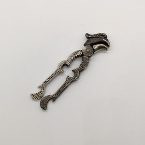 May include: A silver-toned nutcracker shaped like a bird with an ornate design. The nutcracker has a hinged jaw and a bottle opener on each side.