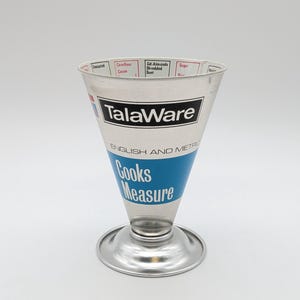 Peut inclure: Une tasse à mesurer TalaWare en métal argenté de forme conique avec une base circulaire. L'extérieur présente des inscriptions en bleu et noir indiquant TalaWare, English and Metric, et Cooks Measure, avec des ingrédients inscrits sur le bord.
