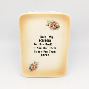 Könnte beinhalten: Cremefarbener rechteckiger Scherenhalter mit floralen Akzenten. Der Halter trägt den Text "I Keep My SCISSORS In This Rack... If You Use Them Please Put Them BACK!" in schwarzer Schrift. Dekorativer Artikel.