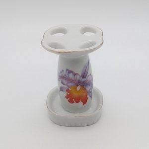 May include: A white ceramic toothbrush holder with a four-hole top and a matching base. The cylindrical body features a painted floral design in shades of purple, orange, and yellow. The edges of the top and base are accented with a thin gold-coloured trim.