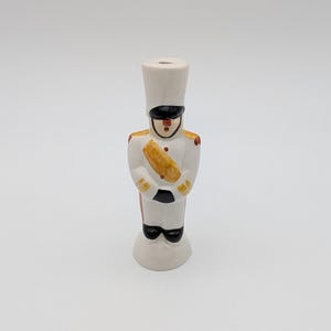Op de afbeelding: Een keramieken beeldje van een speelgoedsoldaat in een wit uniform met zwarte laarzen en een hoge witte hoed. De soldaat heeft een zwart oogmasker, een gouden sjerp en rode accenten. Het beeldje staat rechtop.
