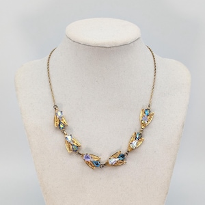 Op de afbeelding: Een vintage goudkleurige ketting met iriserende blauwe en paarse kristallen accenten. De ketting heeft een delicate ketting en sierlijke bladvormige zettingen, die de kleurrijke edelstenen laten zien. De ketting wordt op een mannequin getoond.