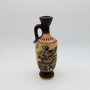 May include: A small, dark brown ceramic vase with a handle and a detailed illustration of figures in black and brown. The vase has a light tan background with decorative patterns near the top.