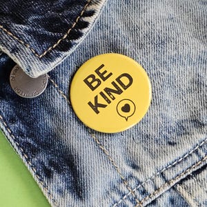 May include: Yellow button with the text "BE KIND" in black letters with a small black heart in a speech bubble. The button is pinned to a blue denim jacket.