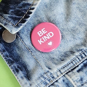May include: Pink button with the text "BE KIND" in white with a small white heart icon. The button is pinned to a blue denim jacket.