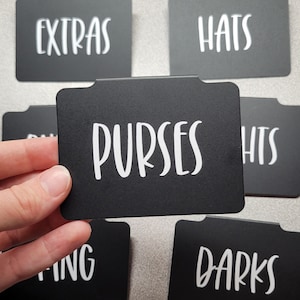 May include: Black metal labels with white text for organizing clothing. The labels read "Extras", "Hats", "Purses", "Darks", and "Hts".