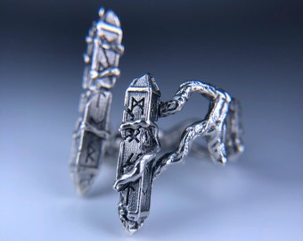 May include: A silver ring with two silver charms. The charms are shaped like pillars with runes carved into them. The ring is designed to look like a vine wrapping around the charms.