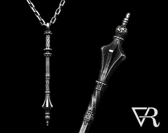 May include: A silver pendant necklace featuring a detailed, ornate design resembling a scepter or staff. The pendant has a pointed tip and a decorative top with intricate details. The necklace is on a silver chain.