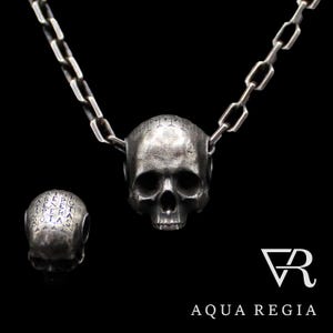 May include: A silver skull pendant necklace with a chain. The skull has a detailed design and is engraved with text. The pendant is attached to a silver chain.