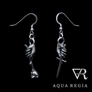 Può includere: Un paio di orecchini d'argento con due mani, una che tiene una rosa e l'altra una spada. Gli orecchini sono su uno sfondo nero con il testo "AQUA REGIA" e un logo "VR" stilizzato.