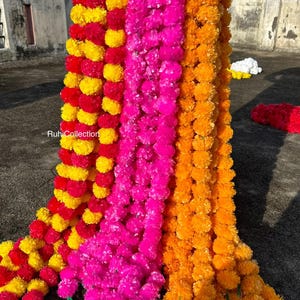 May include: A display of colourful marigold garlands in shades of red, yellow, pink, and orange. The garlands are made of small, round, fluffy flowers and are hanging vertically. The text "RuhiCollection" is visible.