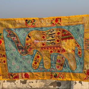 May include: A vibrant textile with an elephant motif. The elephant is yellow with detailed patterns, set against a light blue backdrop. The border is yellow with floral and geometric designs. The textile is held up by unseen hands. The text "Runi collection" is visible.