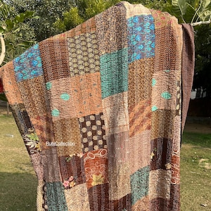 Puede incluir: Un edredón patchwork con una variedad de telas marrones, turquesas y naranjas. El edredón está hecho de cuadrados y rectángulos de diferentes patrones y texturas, con costuras visibles. El edredón está colgado de una rama.