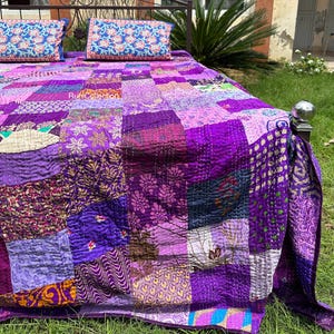 Könnte beinhalten: Eine Patchwork-Tagesdecke aus Stoffquadraten in verschiedenen Violett-, Purpur- und Lavendeltönen mit unterschiedlichen Blumen- und geometrischen Mustern. Die Decke liegt auf einem Metallbettgestell mit zwei passenden rechteckigen Kissen.