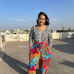May include: A woman wearing a white and black patterned top with a red, blue, and yellow patterned pair of pants. The pants have a drawstring waist and a wide leg.