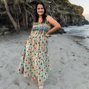 May include: A colorful, patterned maxi dress with a fitted bodice and a flowing skirt. The dress features a mix of floral and geometric designs in shades of green, red, and blue on a cream background. The dress is worn on a beach with palm trees.