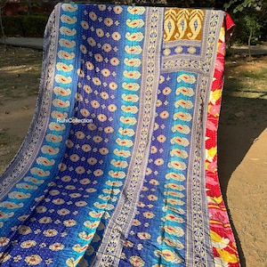 May include: A colorful kantha quilt with a blue, teal, and gold paisley pattern. The quilt is made of recycled saris and has a hand-stitched design. The quilt is folded in half and is lying on a brown surface.