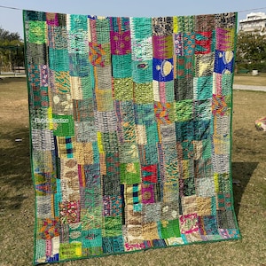 Puede incluir: Un edredón de retazos con una vibrante mezcla de colores, incluyendo verdes, azules y morados. El edredón está hecho de varias piezas de tela cosidas juntas, con un borde verde. El edredón está colgado al aire libre en un tendedero.