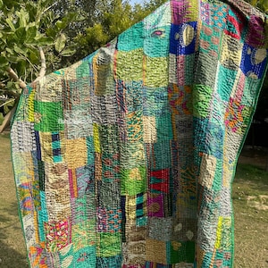 Könnte beinhalten: Ein Patchwork-Quilt mit einer lebendigen Mischung aus Farben wie Grün, Blau, Rot und Gelb. Der Quilt besteht aus verschiedenen Stoffquadraten, die zusammengenäht sind und eine strukturierte Oberfläche bilden. Die Kanten sind mit einem grünen Rand versehen. Der Quilt wird im Freien gezeigt.