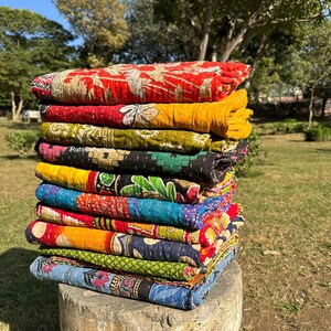May include: A stack of colorful kantha quilts, made from recycled saris, are folded and stacked on a stone pedestal. The quilts are a mix of red, yellow, blue, green, and black.