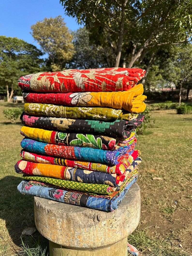 May include: A stack of colorful kantha quilts, made from recycled saris, are folded and stacked on a stone pedestal. The quilts are a mix of red, yellow, blue, green, and black.