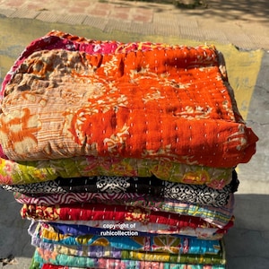 May include: A pile of vibrant, patterned quilts. The top quilt is orange with a floral design, and the others are various colours and patterns. The quilts are hand-stitched with white thread.