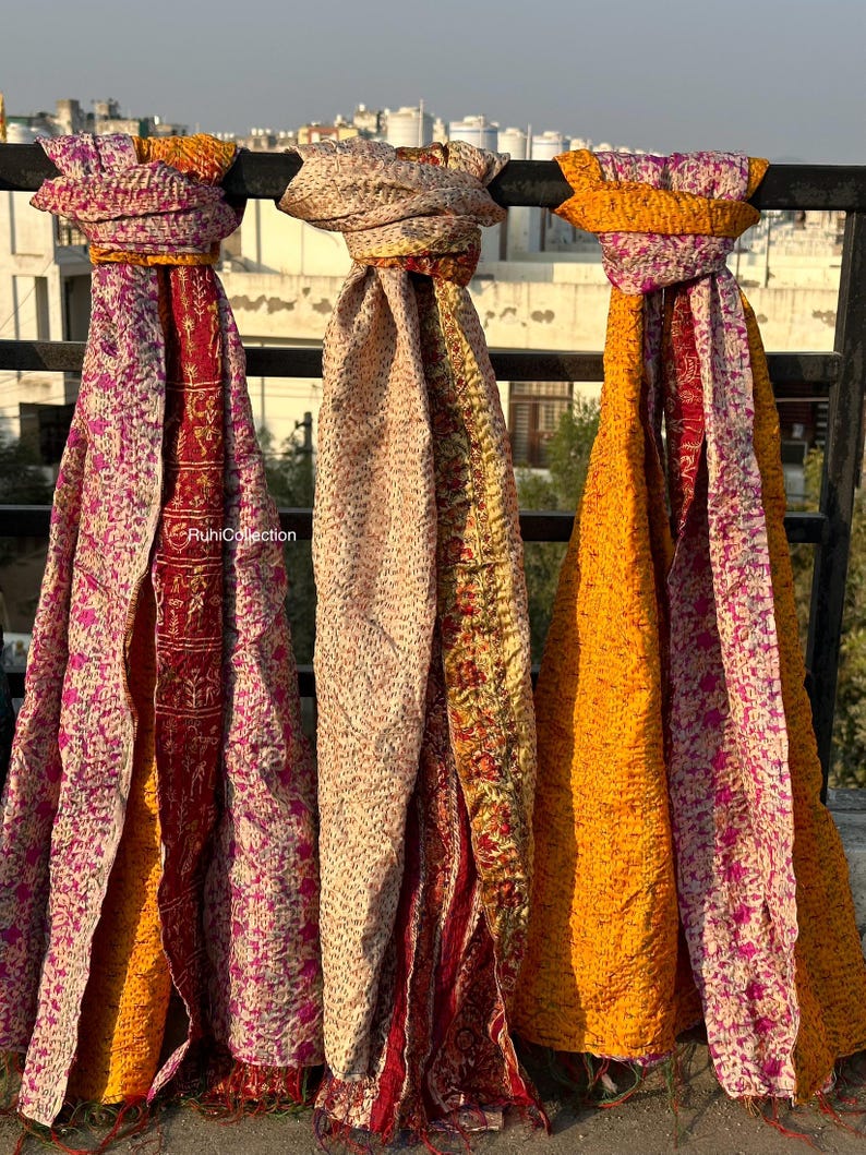 May include: Three colorful silk scarves with a patchwork design. The scarves are hanging on a railing and are made of a variety of fabrics. The scarves are in shades of pink, yellow, and orange.