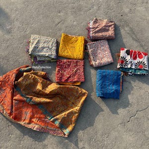 May include: A collection of eight colorful kantha quilts, a traditional Indian textile, in various patterns and colors, including red, blue, yellow, and white. The quilts are folded and arranged on a gray concrete surface. The text "RuhiCollection" is visible in the lower left corner of the image.