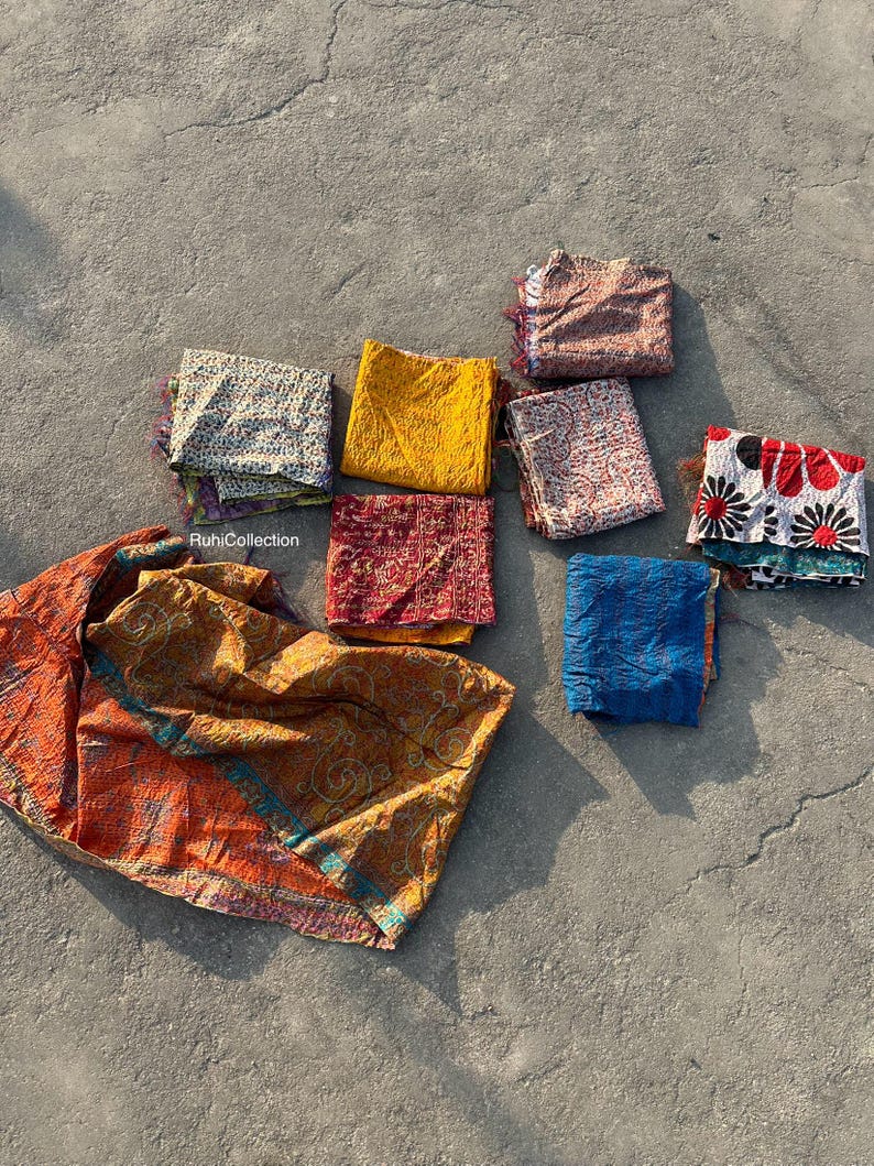 May include: A collection of eight colorful kantha quilts, a traditional Indian textile, in various patterns and colors, including red, blue, yellow, and white. The quilts are folded and arranged on a gray concrete surface. The text "RuhiCollection" is visible in the lower left corner of the image.
