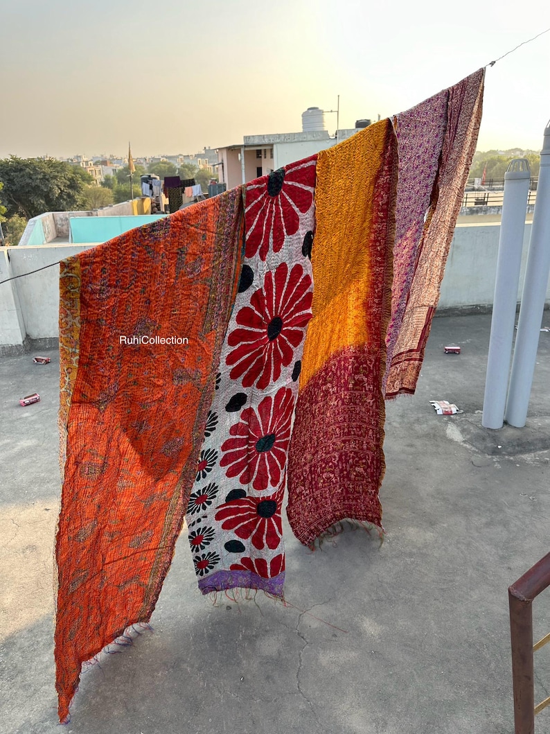 May include: Four colorful kantha quilts with floral patterns hang on a clothesline. The quilts are made of recycled saris and are a popular Indian craft. The quilts are in shades of orange, red, yellow, and purple.
