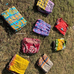 May include: Eight colorful kantha quilts folded and laid out on a green grassy surface. The quilts are made of recycled saris and feature a variety of patterns and colors, including blue, yellow, red, and purple.