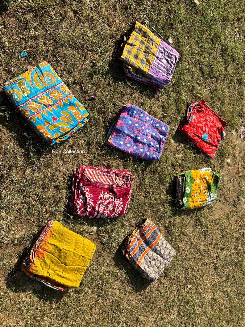 May include: Eight colorful kantha quilts folded and laid out on a green grassy surface. The quilts are made of recycled saris and feature a variety of patterns and colors, including blue, yellow, red, and purple.
