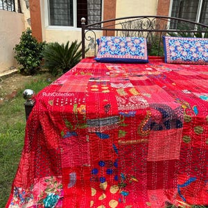 May include: A red kantha quilt with a patchwork design featuring various colours and patterns. The quilt is laid out on a bed with two blue floral patterned pillowcases. The text "RuhCollection" is visible in the lower left corner of the image.