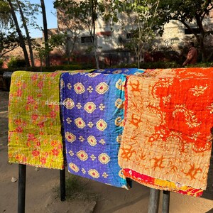 May include: Three kantha quilts hanging on a clothesline. The quilts are made of colorful fabric with intricate patterns. The quilt on the left is green and yellow, the middle quilt is blue and gold, and the quilt on the right is orange and red.