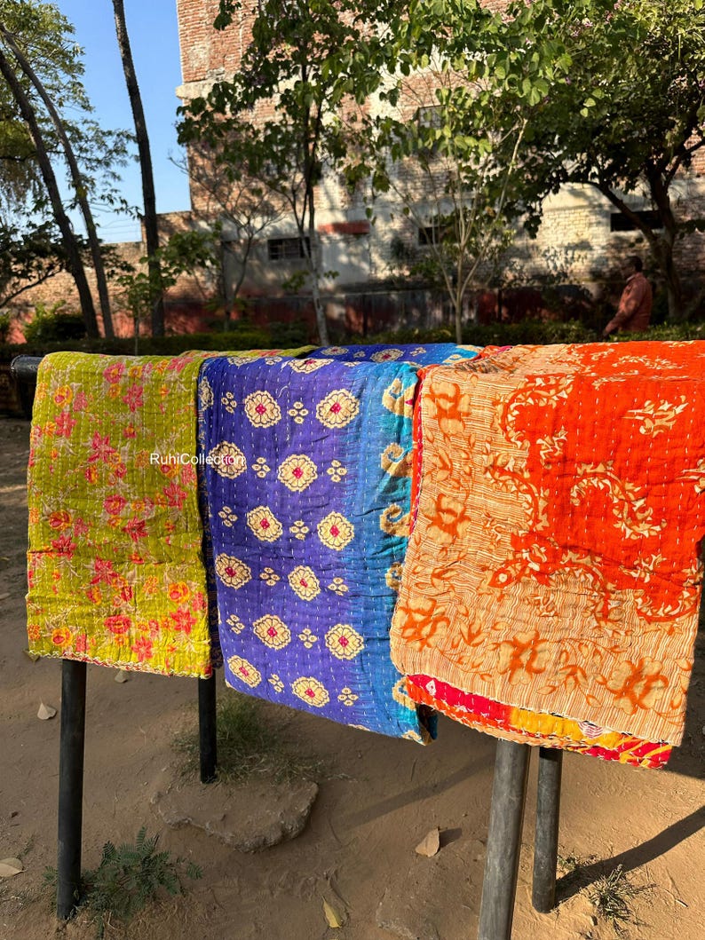 May include: Three kantha quilts hanging on a clothesline. The quilts are made of colorful fabric with intricate patterns. The quilt on the left is green and yellow, the middle quilt is blue and gold, and the quilt on the right is orange and red.