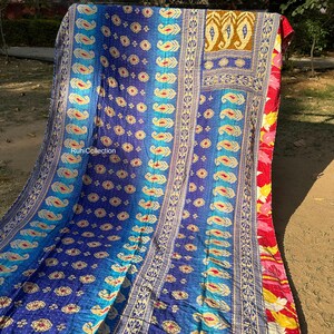 May include: A colorful kantha quilt with a blue, teal, and gold paisley pattern. The quilt is made of recycled saris and has a hand-stitched design. The quilt is folded in half and is lying on a brown surface.