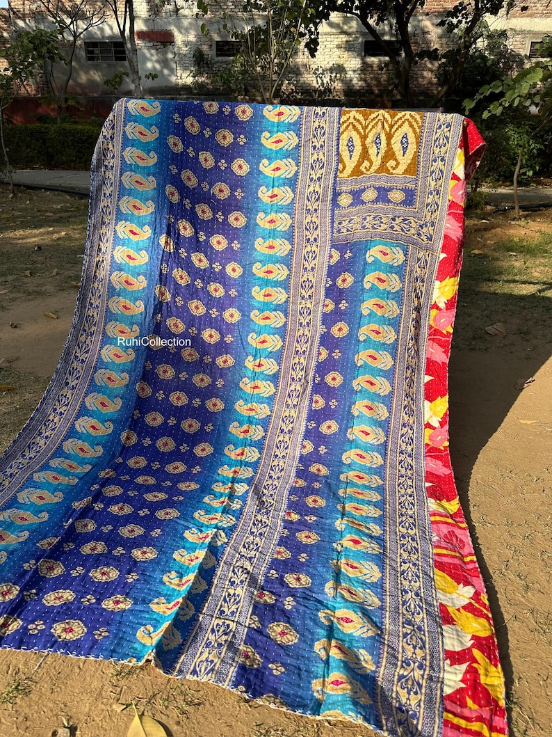May include: A colorful kantha quilt with a blue, teal, and gold paisley pattern. The quilt is made of recycled saris and has a hand-stitched design. The quilt is folded in half and is lying on a brown surface.