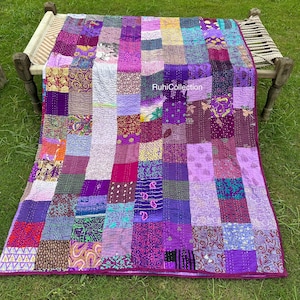 May include: A patchwork quilt in shades of purple, lavender, and magenta, with a variety of patterned fabric squares. The quilt is draped over a wooden frame on a grassy surface. The text "RuhiCollection" is visible.