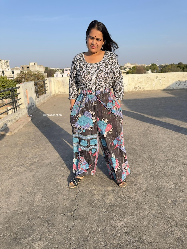 May include: A woman wearing a black and white patterned top with a floral print jumpsuit. The jumpsuit has a drawstring waist and wide legs.