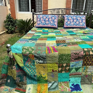 Könnte beinhalten: Eine Patchwork-Tagesdecke aus verschiedenen grünen, türkisfarbenen und gelben Stoffquadraten mit unterschiedlichen Mustern und sichtbaren Nähten. Zwei rechteckige Kissen mit blauem Hintergrund und rot-weißem Blumenmuster liegen auf dem Bett.