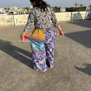 May include: A woman wearing a white and black patterned top with a brown belt and colorful patchwork pants. The pants have a blue, yellow, and turquoise pattern with floral designs.