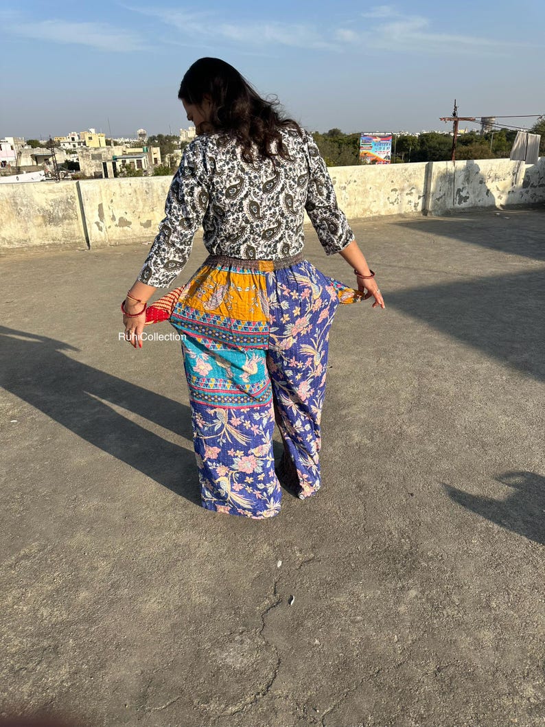 May include: A woman wearing a white and black patterned top with a brown belt and colorful patchwork pants. The pants have a blue, yellow, and turquoise pattern with floral designs.