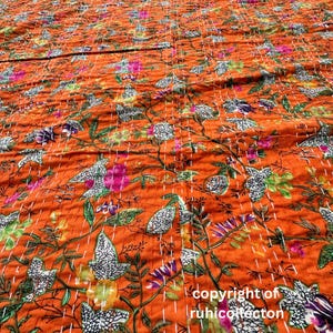 May include: Orange kantha quilt with a floral pattern. The quilt is hand-stitched and features a variety of colors, including green, pink, and white. The quilt is made of cotton and is perfect for adding a touch of bohemian style to your home.
