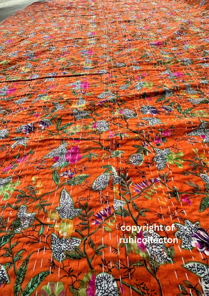 May include: Orange kantha quilt with a floral pattern. The quilt is hand-stitched and features a variety of colors, including green, pink, and white. The quilt is made of cotton and is perfect for adding a touch of bohemian style to your home.
