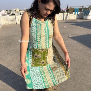 May include: A sleeveless apron with a patchwork design in shades of green, cream, and gold. The apron features a large pocket with a floral pattern and is worn over patterned pants. The apron is made of a soft, quilted fabric.
