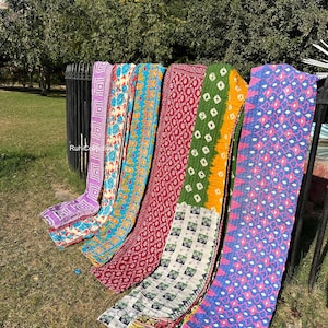 May include: Six colorful kantha quilts with various patterns and designs, including floral, geometric, and paisley motifs. The quilts are arranged in a row, hanging from a metal fence. The quilts are made of recycled saris and are a popular choice for home decor and gifts.