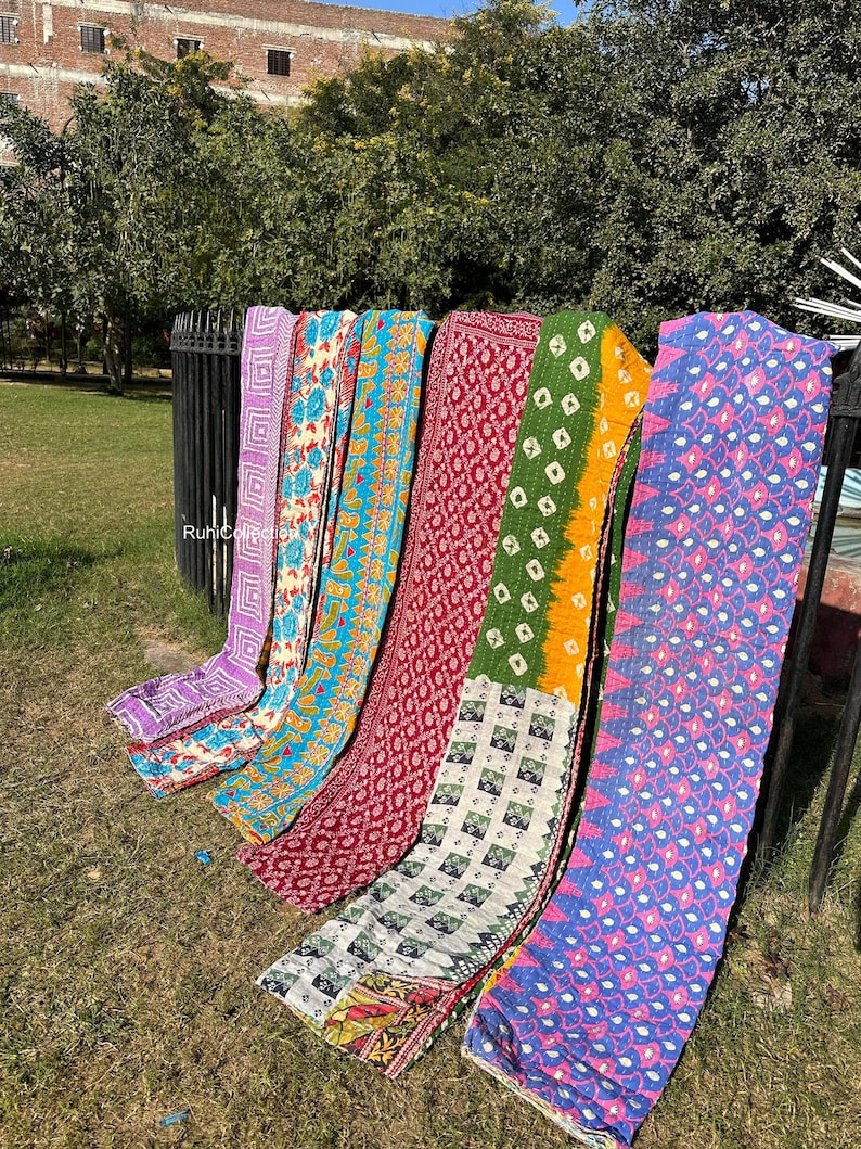 May include: Six colorful kantha quilts with various patterns and designs, including floral, geometric, and paisley motifs. The quilts are arranged in a row, hanging from a metal fence. The quilts are made of recycled saris and are a popular choice for home decor and gifts.
