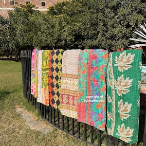 May include: A row of colorful, patterned quilts hanging on a black metal fence. The quilts feature various designs, including floral, geometric, and leaf patterns, in shades of pink, green, orange, and yellow. The background includes trees and a building.