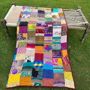 以下が含まれることがあります： 紫、黄色、青、赤など、さまざまな色と模様が混ざり合ったパッチワークキルト。 キルトは木製のベッドフレームに掛けられており、長方形のデザインと縫製構造を示しています。 キルトには濃い赤色の縁取りがあります。