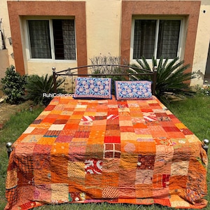 May include: A patchwork quilt with a mix of orange, red, and brown squares. The quilt is on a bed frame with two blue floral patterned pillows. The bed is in front of a house with two windows.